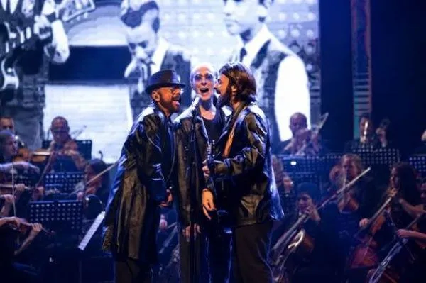 Três cantores de jaqueta de couro cantam ao microfone diante de uma orquestra