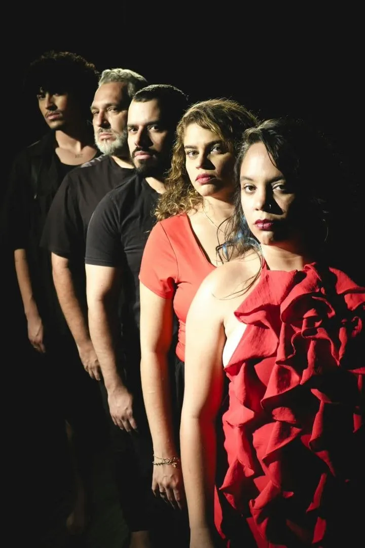 Cinco atores em fila, de perfil, a primeira com vestido vermelho de babados