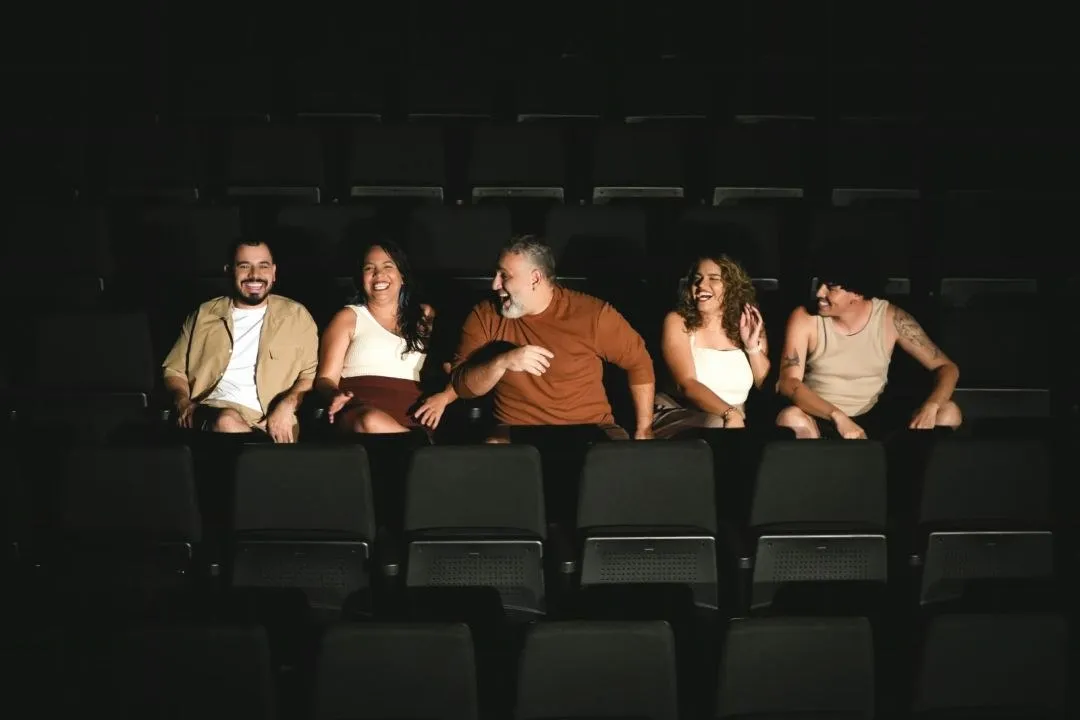 Cinco atores sentados lado a lado na plateia escura de um teatro