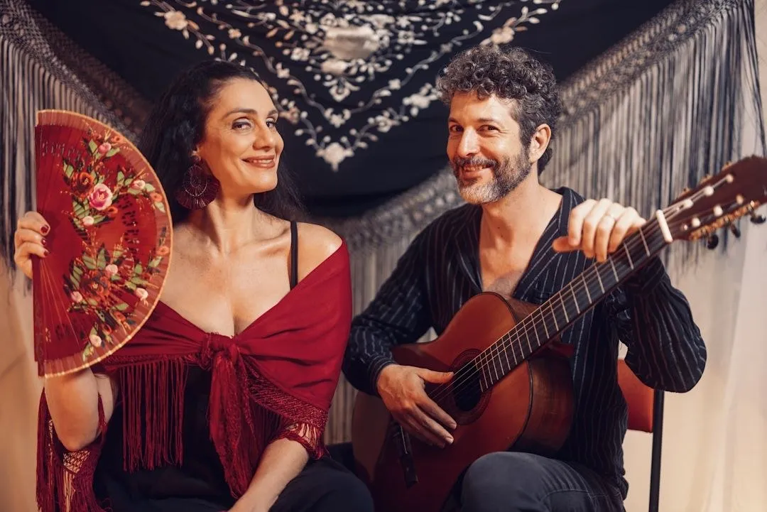 Fernanda Cabral, com leque vermelho, ao lado de violonista sentado com o instrumento