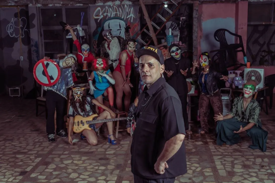 Elenco de Danúbio Azul, com máscaras de caveira e figurinos coloridos, em frente a um muro grafitado