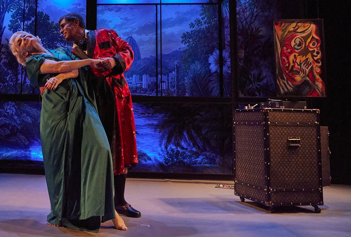 Atriz de vestido verde se inclina nos braços de ator de roupão vermelho, em cena de teatro