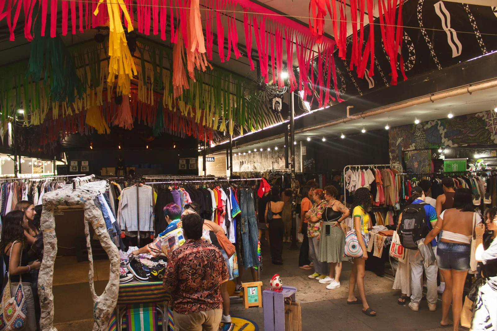 Público entre araras de roupas sob bandeirolas coloridas, em feira de moda circular