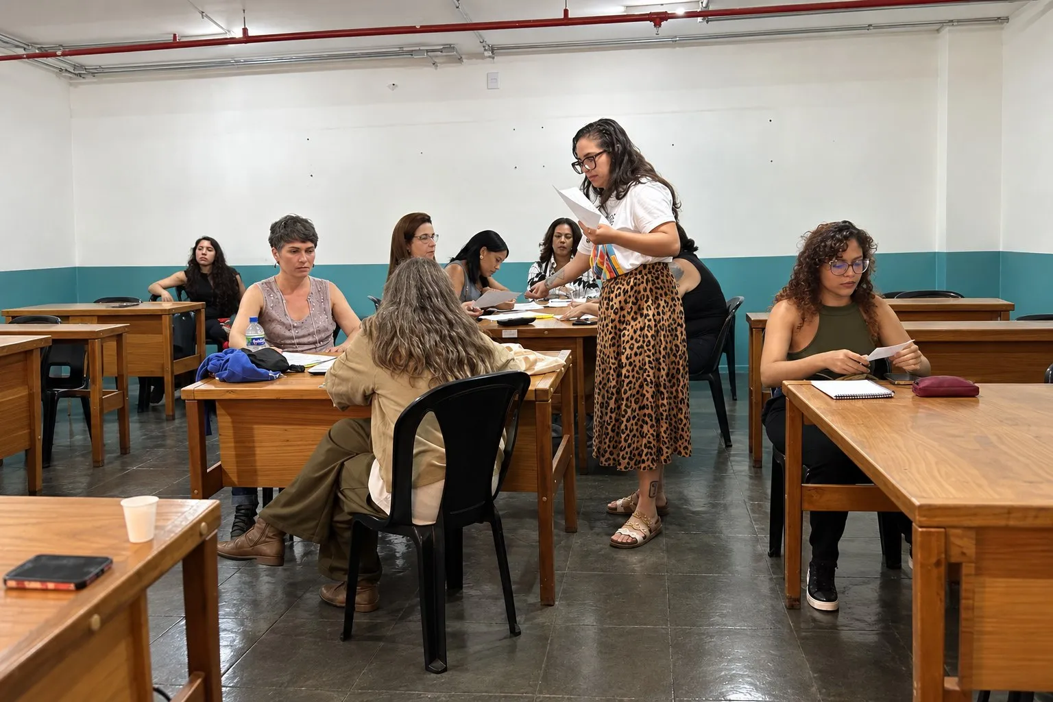 Instrutora em pé distribui papéis a mulheres sentadas em carteiras, em sala de aula